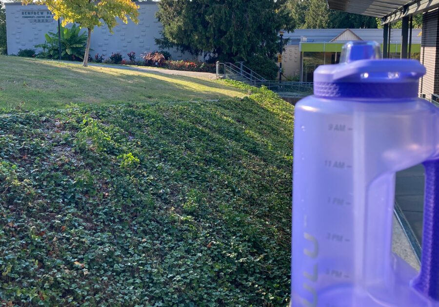 バンクーバーのコミュニティセンター外の芝生を背景に置かれた、目盛り付きの紫色の大きな水筒。節約と健康管理のために飲み物を持ち歩く日常の風景。